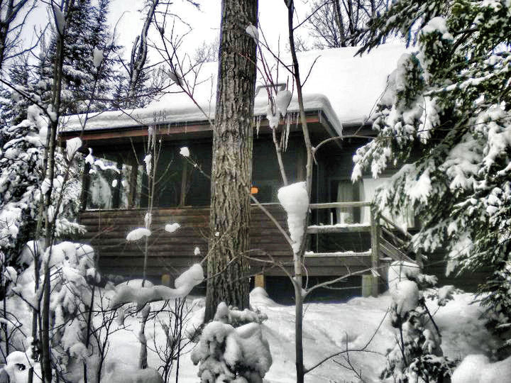 Outside of a log cabin in the winter with trees covered in snow and icicles on roof