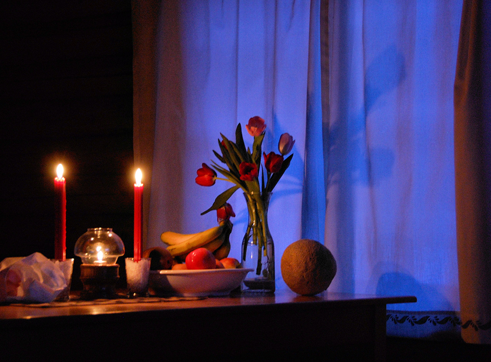 Table with fruit, vase with red flowers, and two lit red candles in the dusk