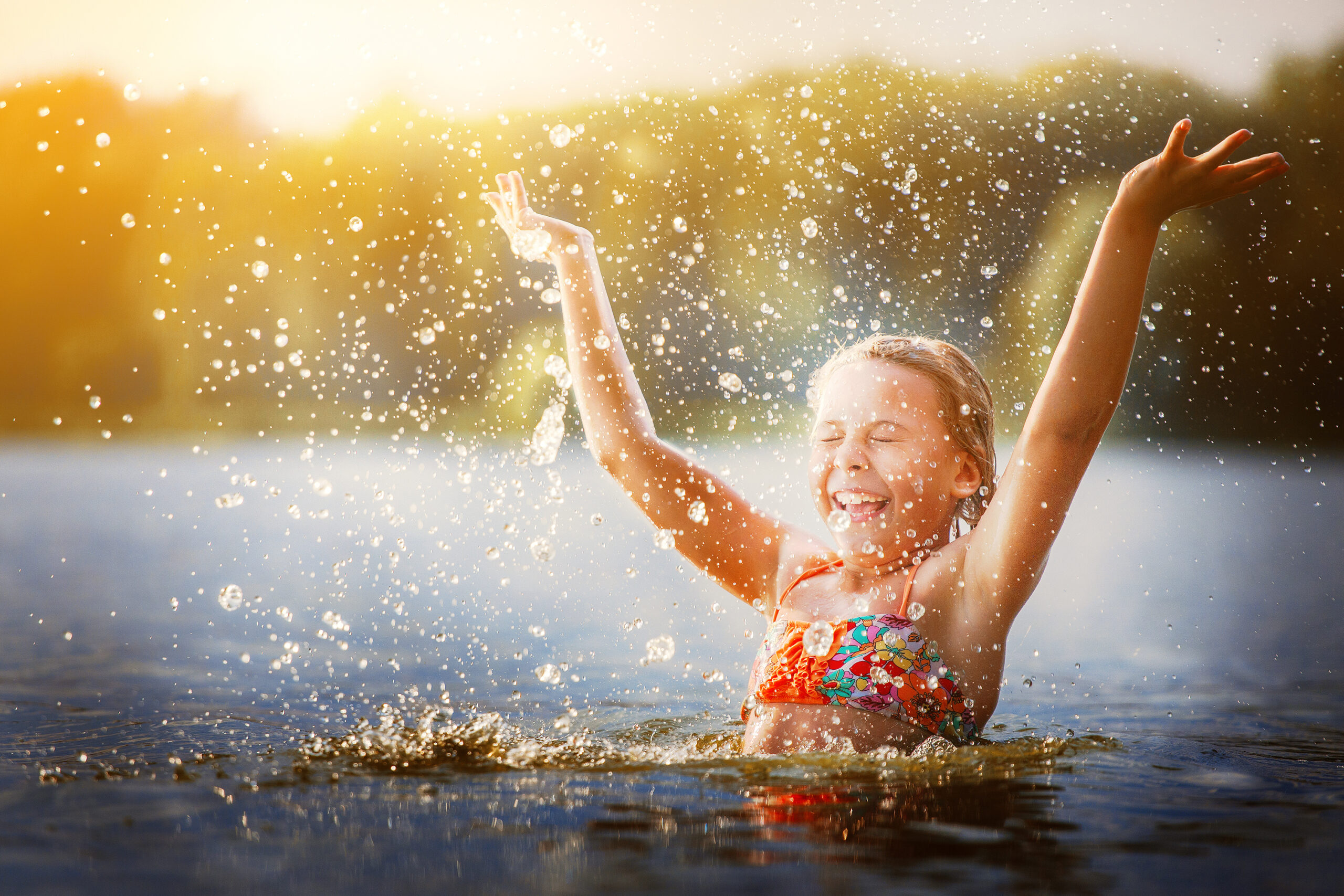 A girl with blond hair playing in the river, she raises her hands up in the water and splashes water drops